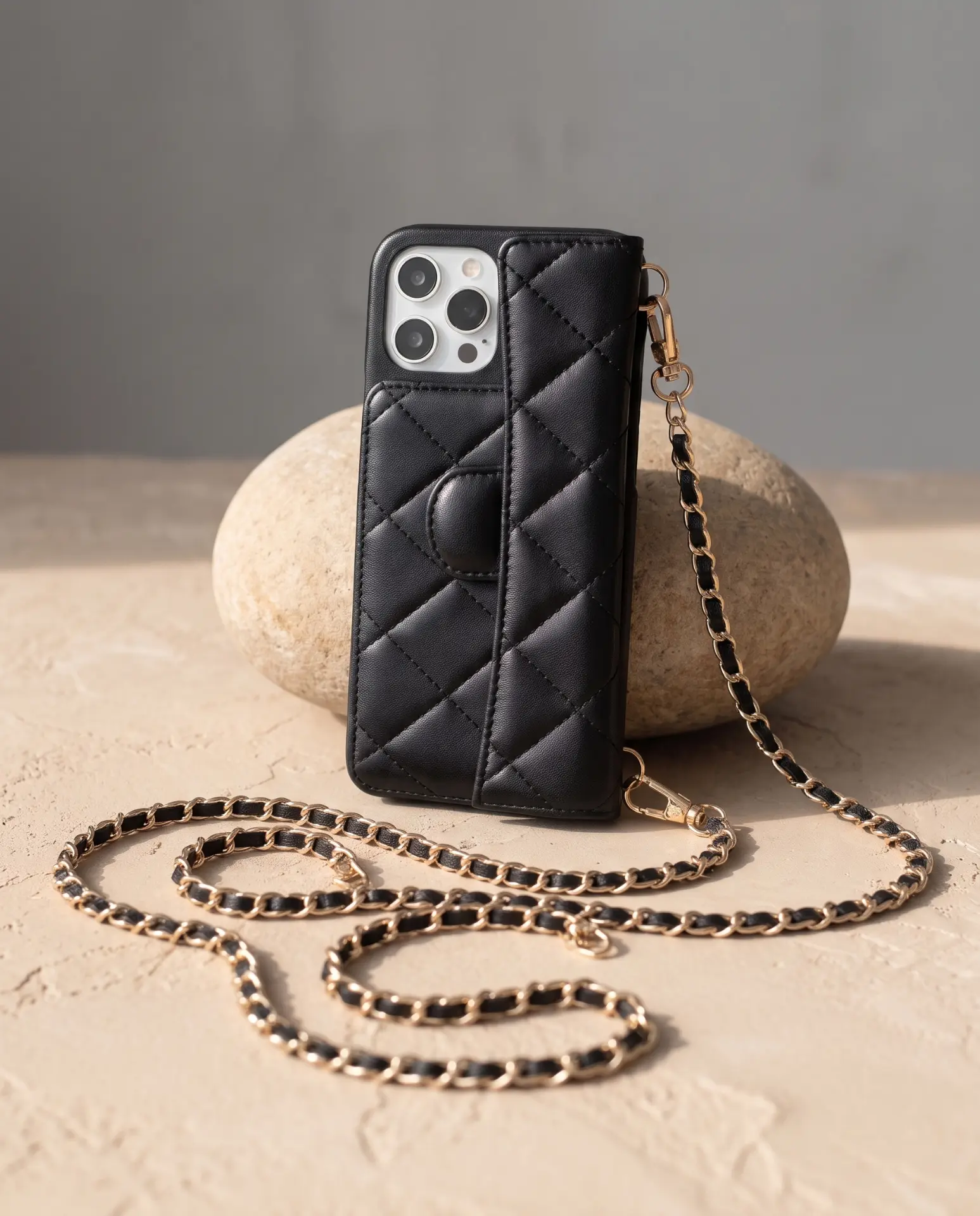 Midnight Black phone purse with a full-length pocket, propped upright against a tan river stone with its gold chain winding across the sand
