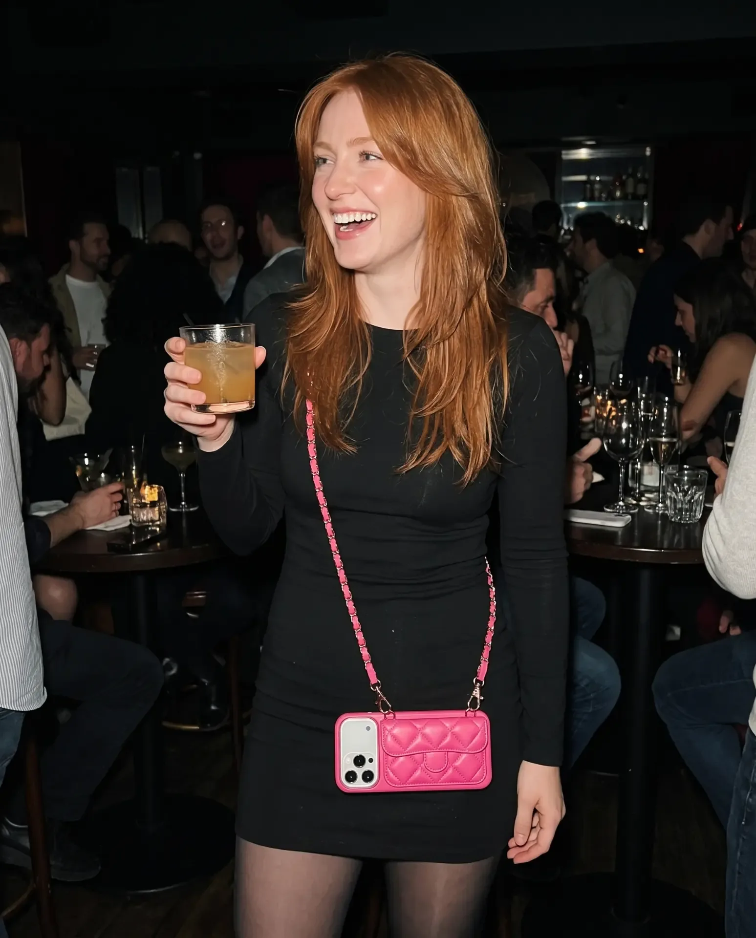 Powder Pink phone purse with a full-length pocket worn crossbody in a dim cocktail lounge, held against a black dress with a drink in hand