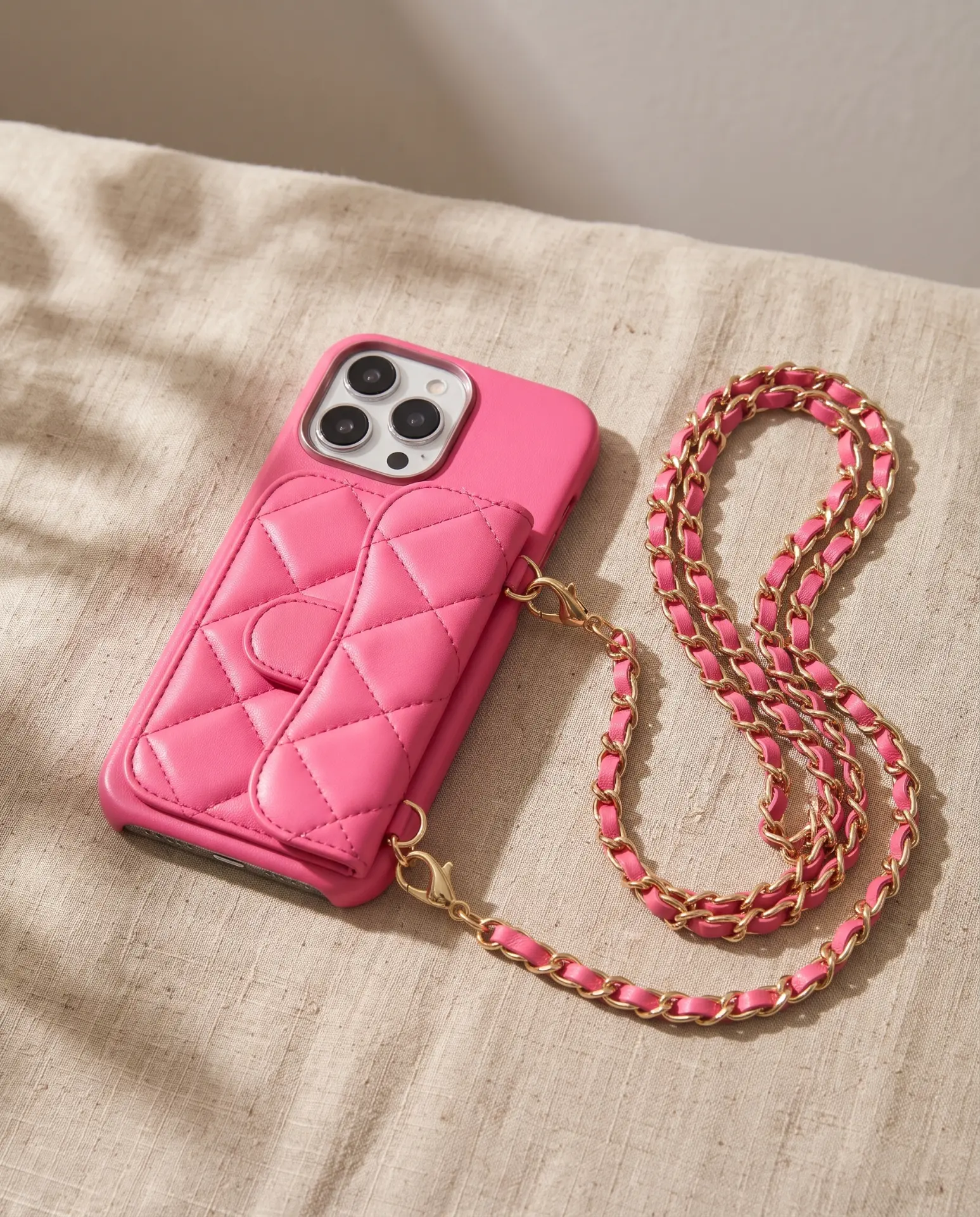Powder Pink phone purse with a short pocket, resting at an angle on woven linen with its gold chain coiled beside it