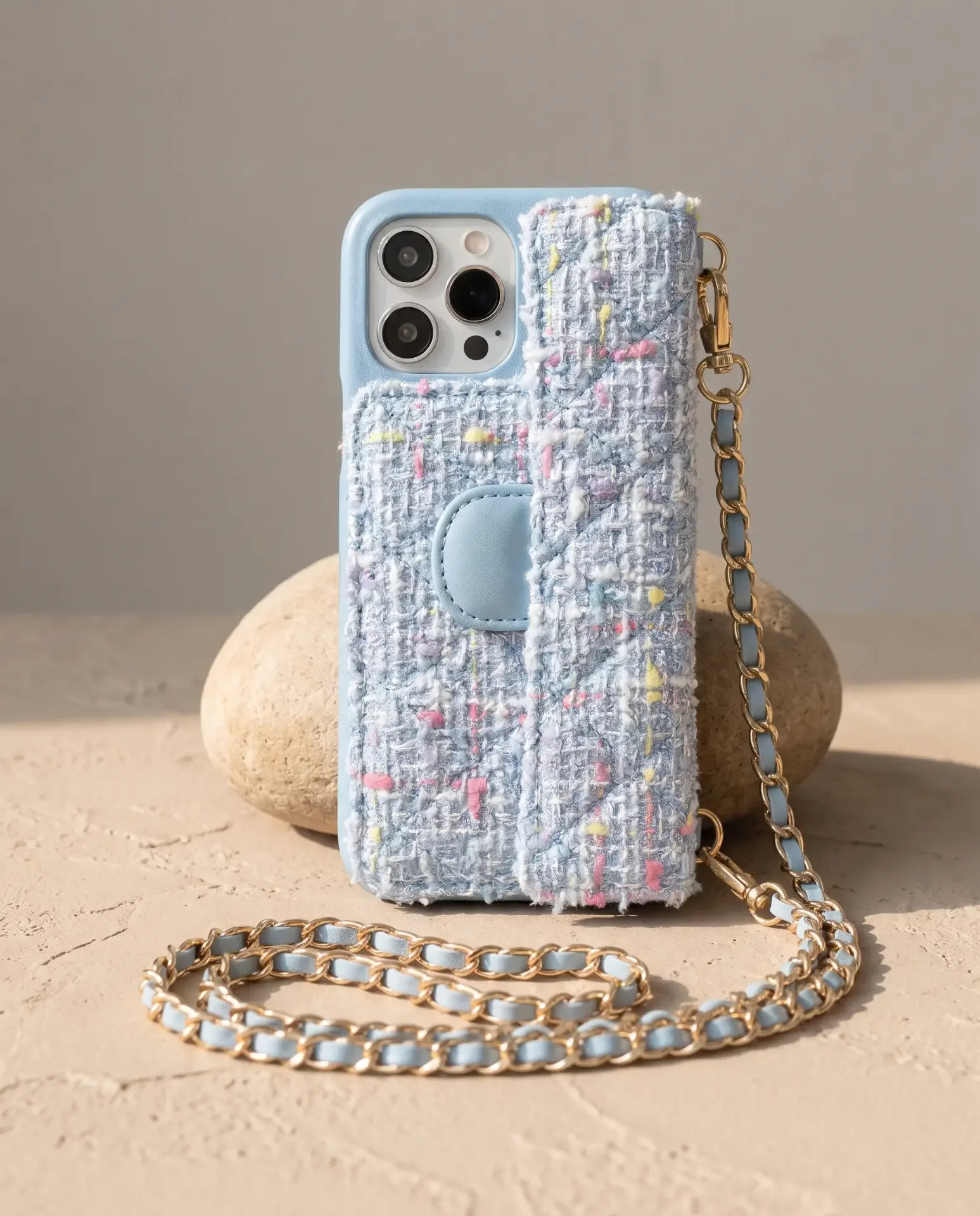 Sky Blue Tweed phone purse with a full-length pocket, propped upright against a smooth stone with its gold chain looped in front
