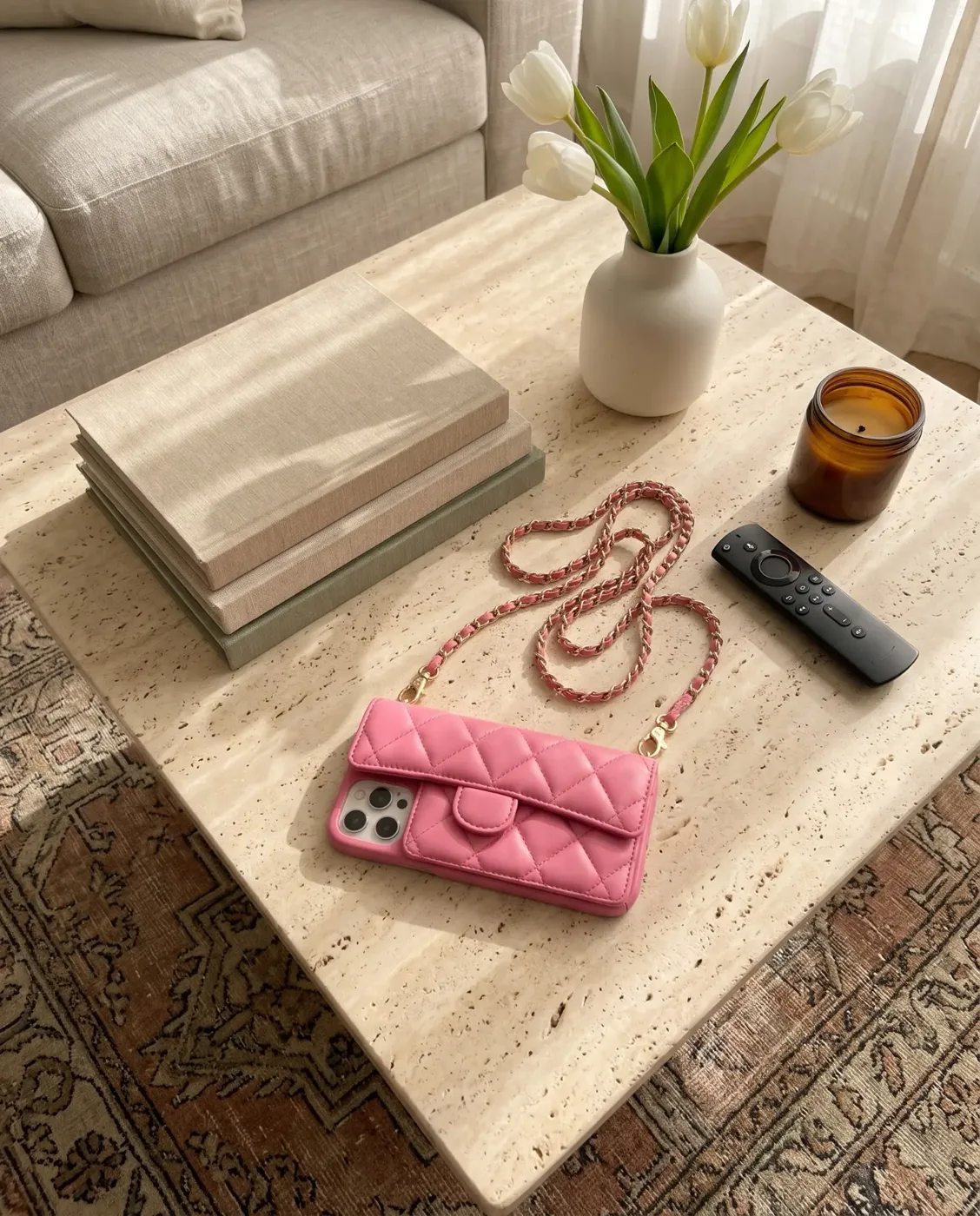 Powder Pink phone purse with a full-length pocket, on a travertine coffee table with stacked books, white tulips and an amber candle