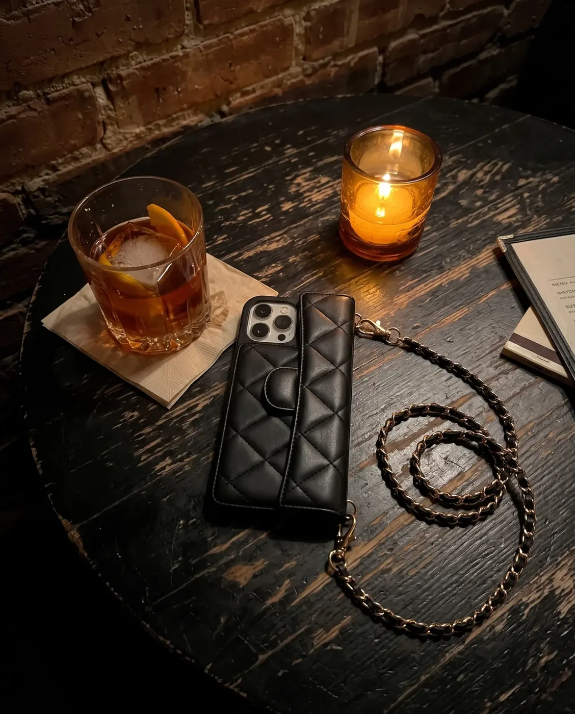 Midnight Black phone purse with a full-length pocket on a dark wooden bar table beside a cocktail and a lit candle, its gold chain coiled in front