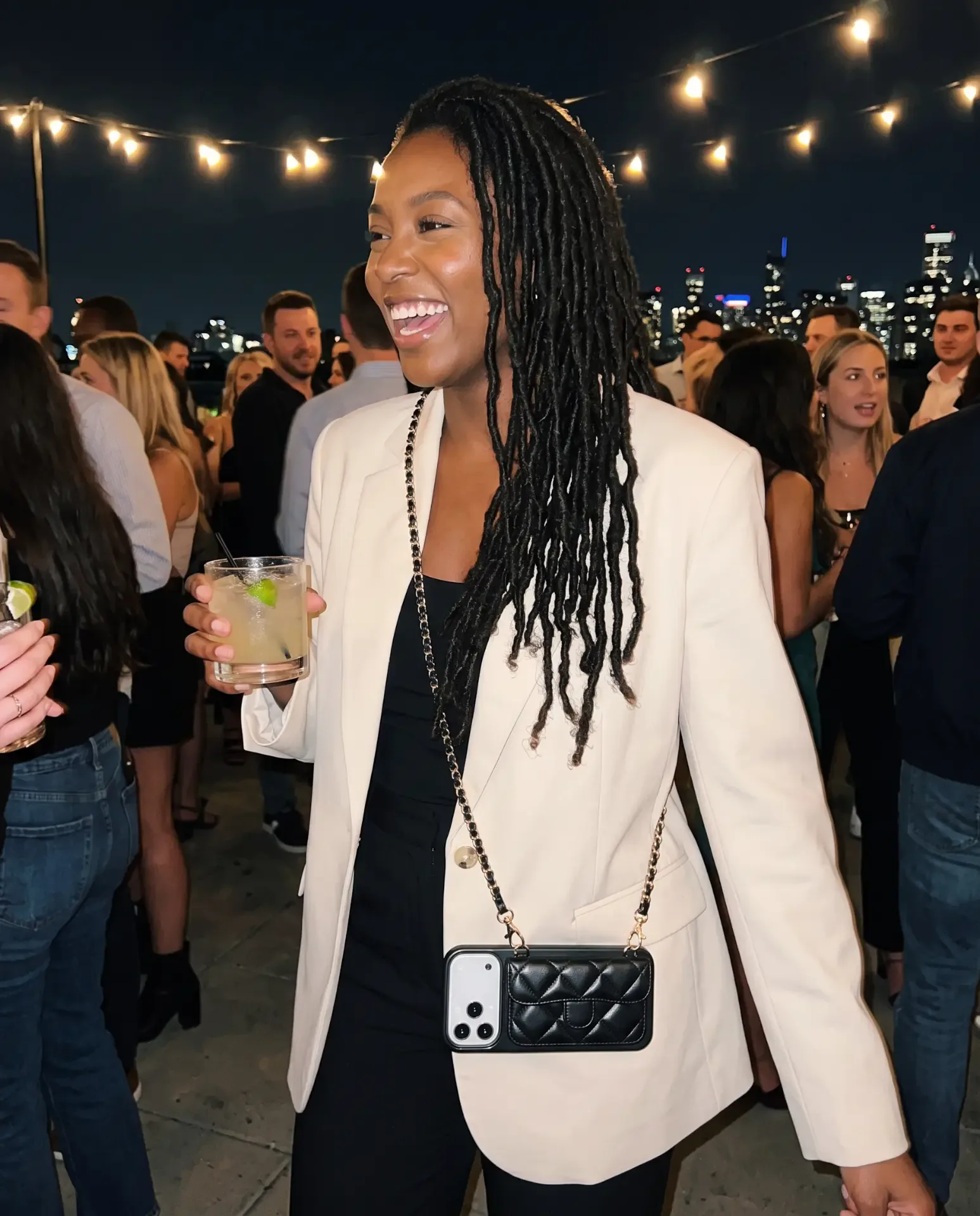 Midnight Black phone purse with a short pocket held at a nighttime rooftop party, string lights, guests and the city skyline behind