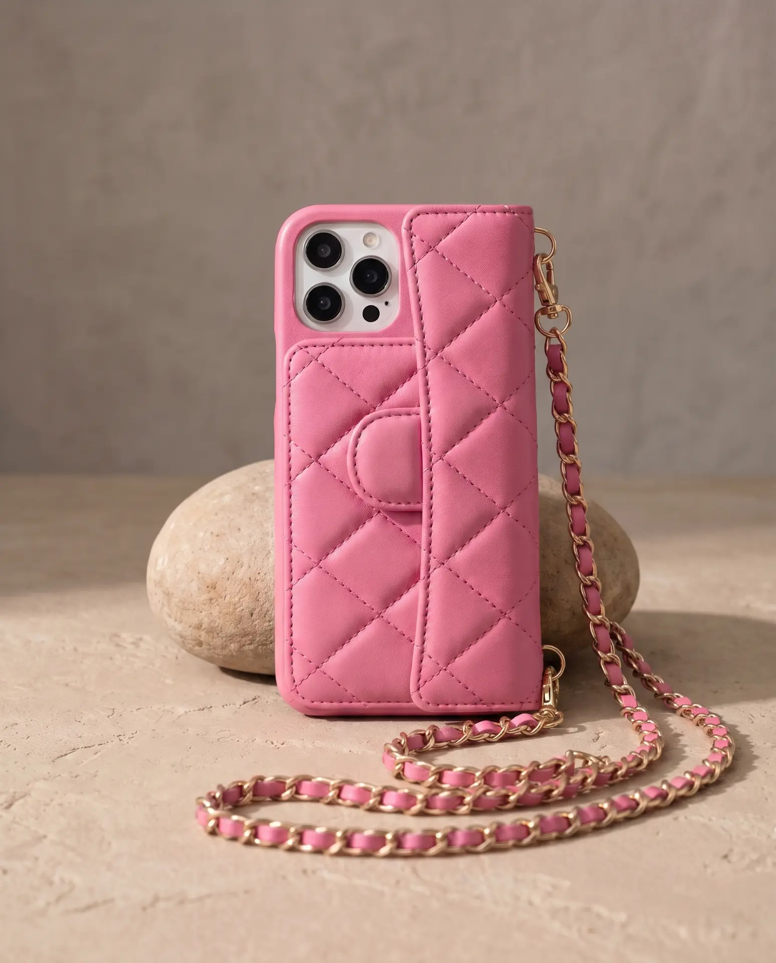 Powder Pink phone purse with a full-length pocket, propped upright against a smooth beige stone with its gold chain pooling in front