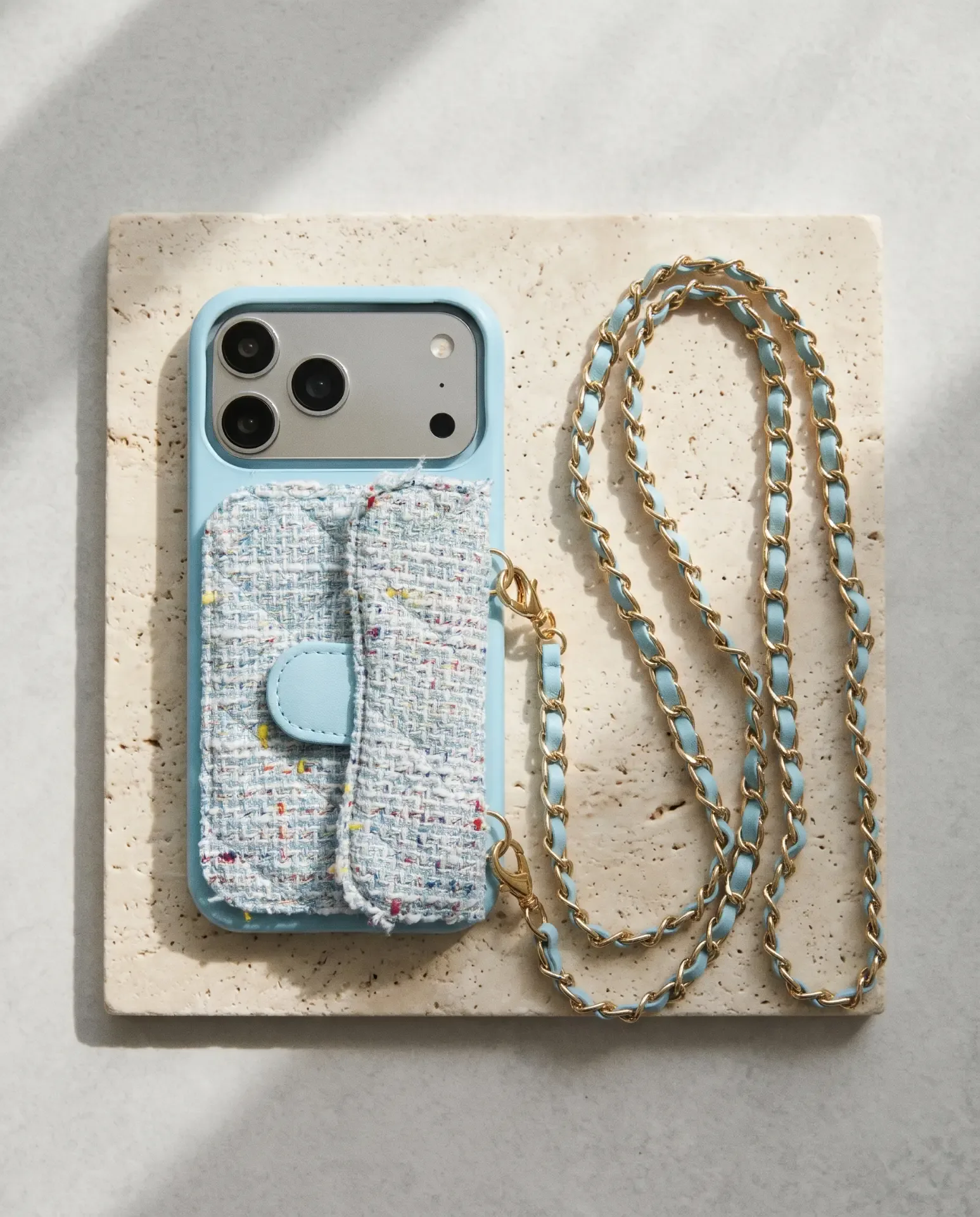 Sky Blue Tweed phone purse with a short pocket, laid flat on a travertine tile with its gold chain looping beside it