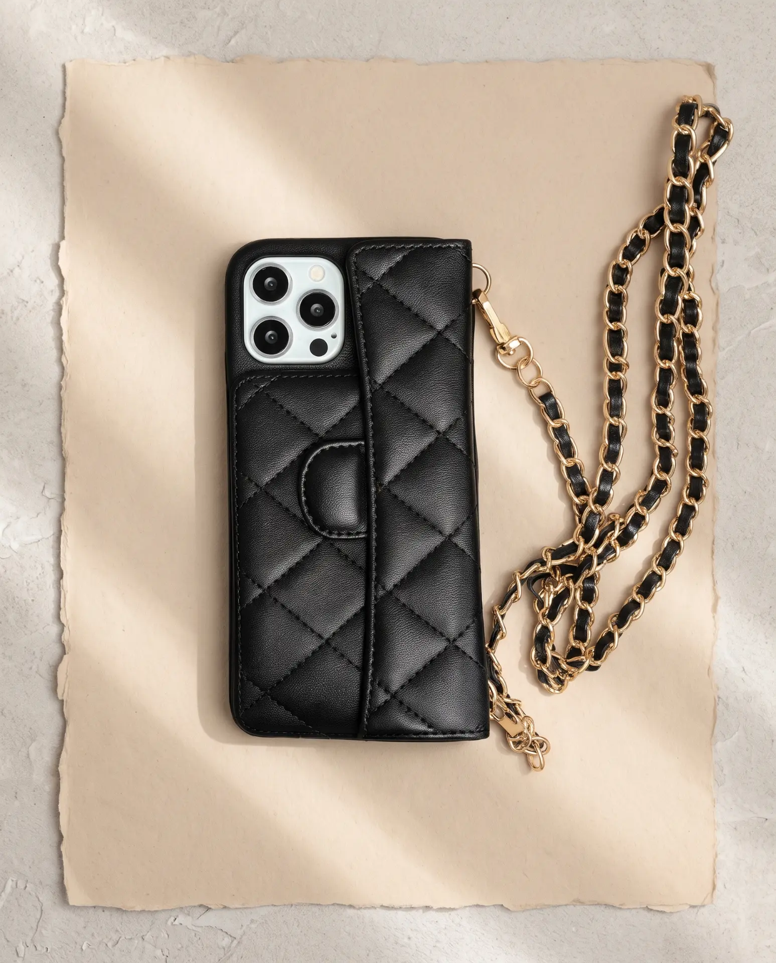 Midnight Black phone purse with a full-length pocket, laid flat on a deckle-edged sheet of paper with its gold chain folded beside it