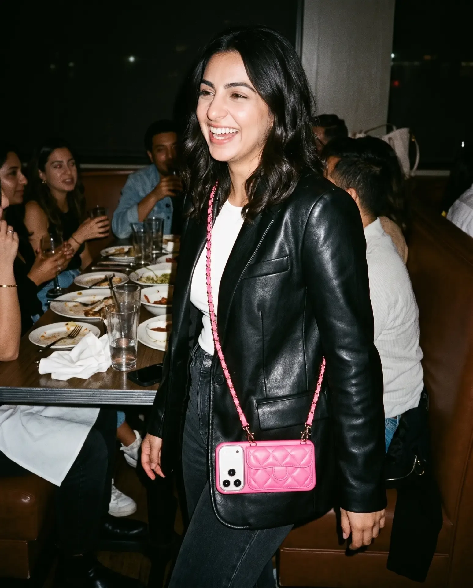 Powder Pink phone purse with a full-length pocket worn crossbody at a dinner table in a busy restaurant booth, plates, wine and friends behind
